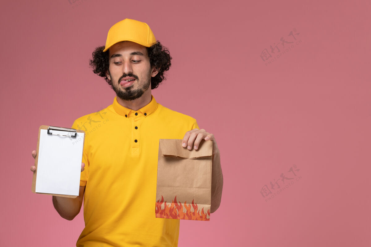 男正面图身穿黄色制服的男快递员拿着食品包和笔记本放在粉红色的墙上查看工人制服