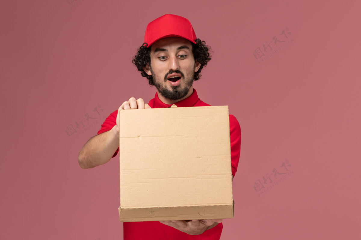 持有正面图身穿红色衬衫和披风的男快递员拿着送货食品箱 在浅粉色的墙上打开正面信封容器