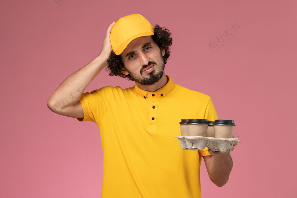 思考正面图穿着黄色制服的男信使拿着棕色的咖啡杯在粉红色的墙上思考快递员棕色咖啡