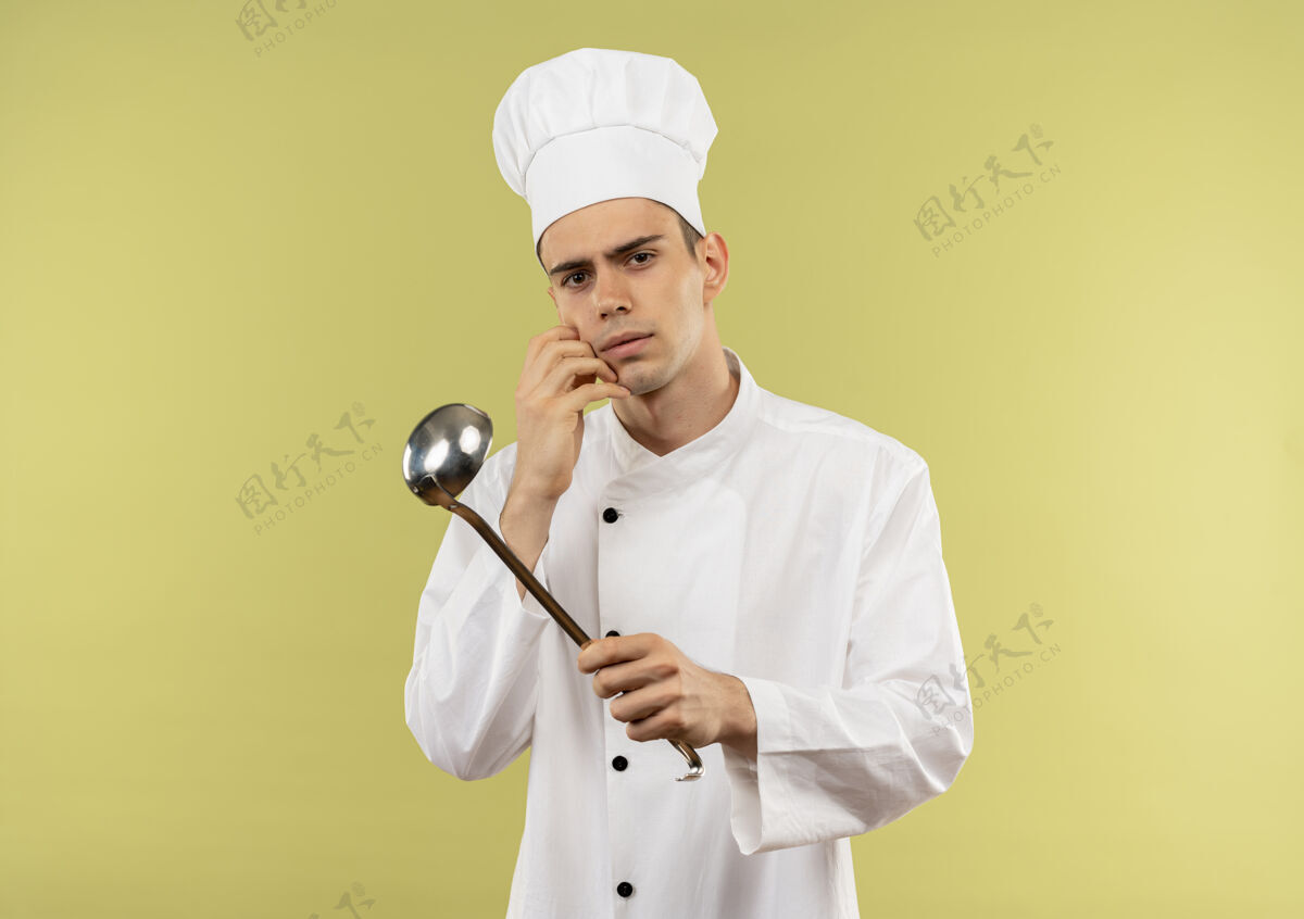 厨师困惑的年轻男厨师穿着厨师制服拿着勺子把手放在面颊上隔离的绿色墙壁与复制空间手拿着脸颊
