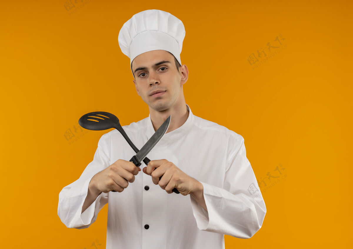 刀年轻的男酷穿着厨师制服 手里拿着勺子和刀 站在隔离的黄色墙壁上厨师看手