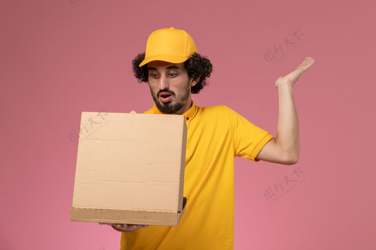 工人正面图：身穿黄色制服的男性快递员手持并打开浅粉色墙上的食品递送箱男快递员持有制服