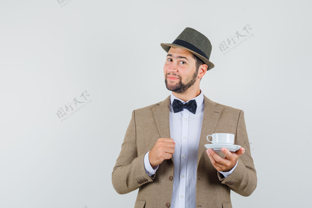 男人年轻人穿着西装 戴着帽子 手里拿着白色的茶杯和茶碟 看上去很开朗 正对着窗外成人年轻看