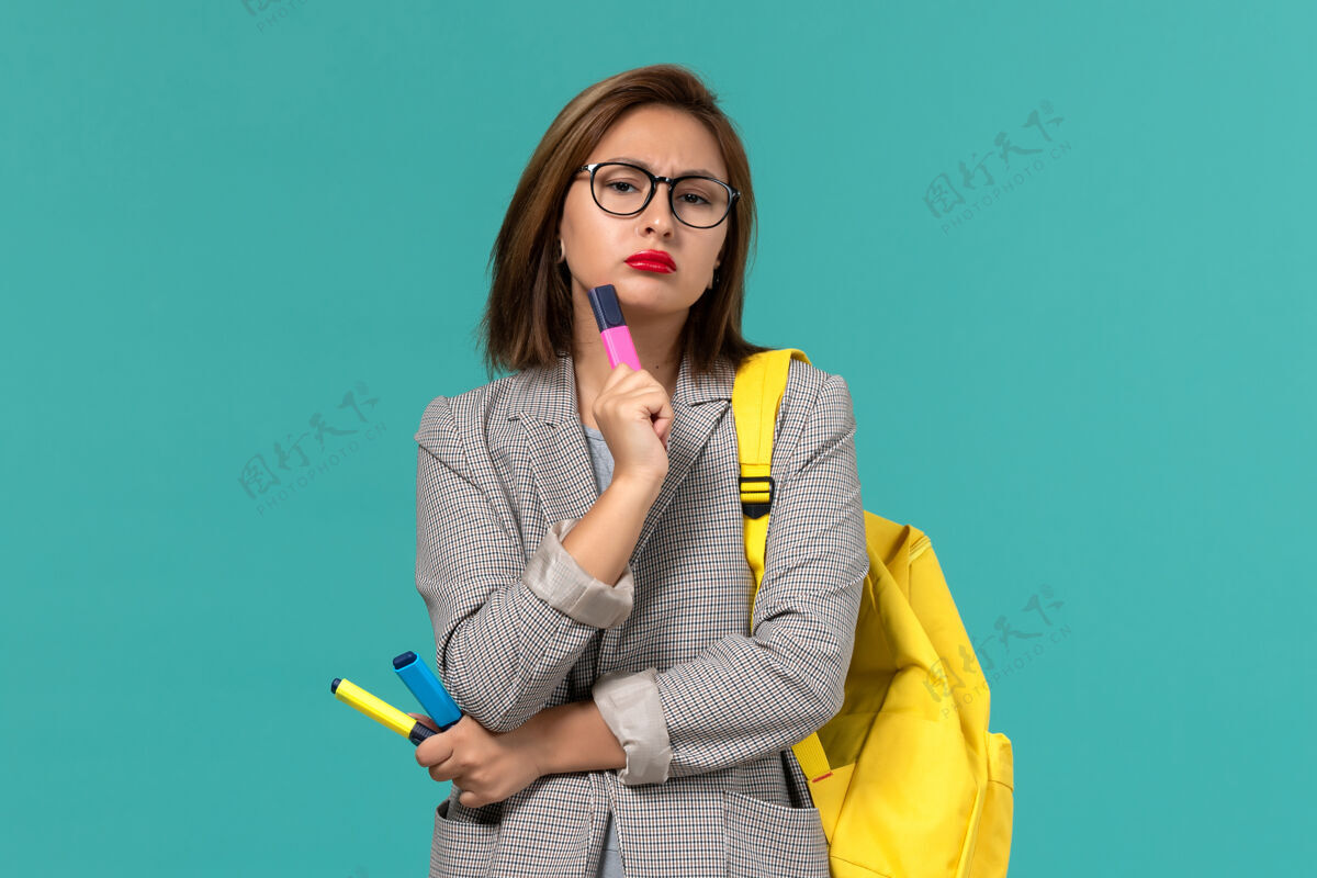 大学身穿灰色夹克 背着黄色背包 拿着毛毡笔 在淡蓝色墙上思考的女学生的正视图人保持教训