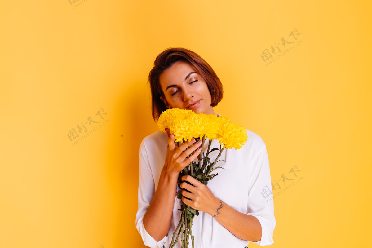 华丽摄影棚拍摄的黄色背景快乐的白人妇女短发穿着休闲服白衬衫和牛仔裤手持一束黄色紫苑微笑看可爱