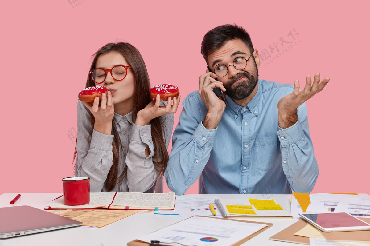 男性快乐女人的照片享受着咖啡休息 吃着美味的甜甜圈 和通过手机聊天的团友一起学习 有着茫然的表情文书工作同事眼镜