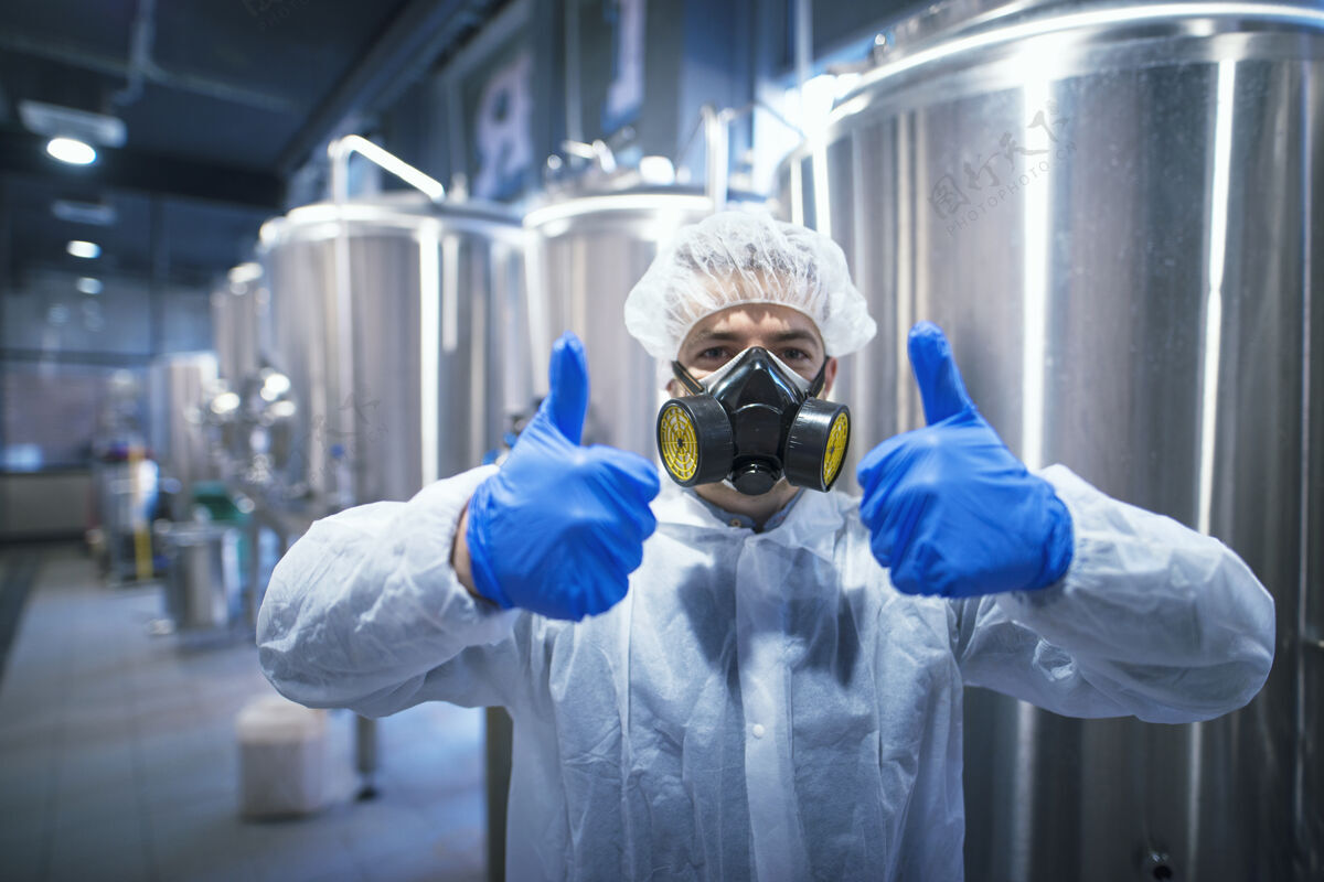 化学家专业技术专家穿着白色防护服 戴着发网 口罩和手套 竖起大拇指工厂检验设备