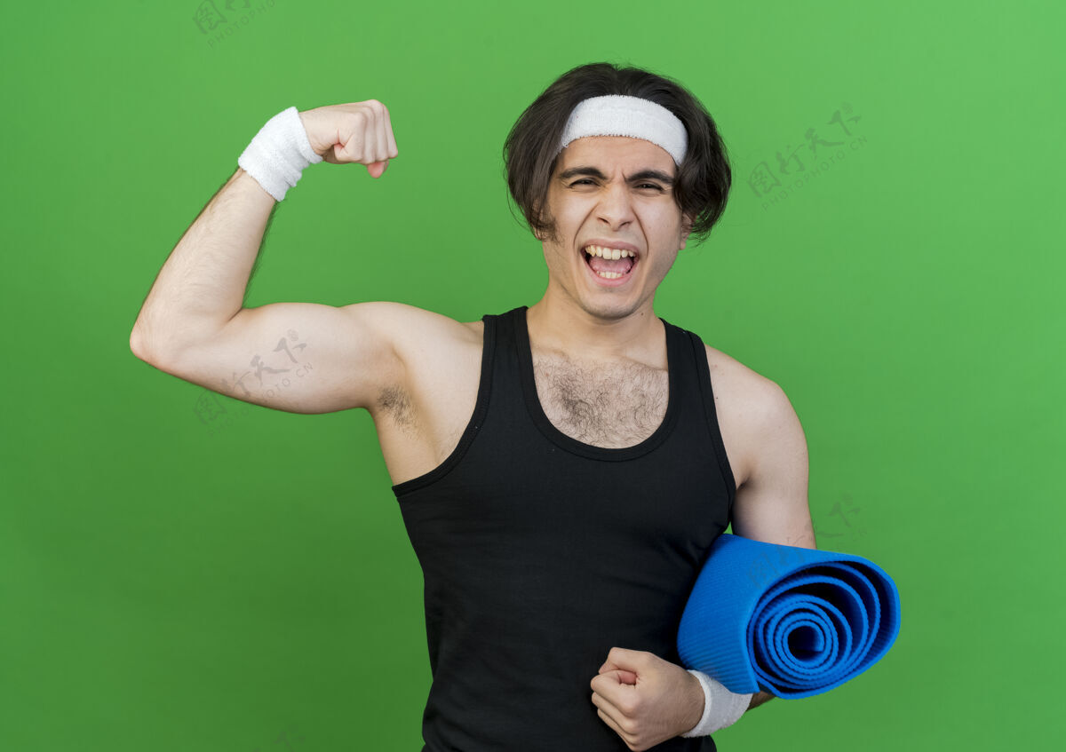 运动装年轻的运动型男子穿着运动服 头戴瑜伽垫举起拳头紧张和自信站着绿色垫子