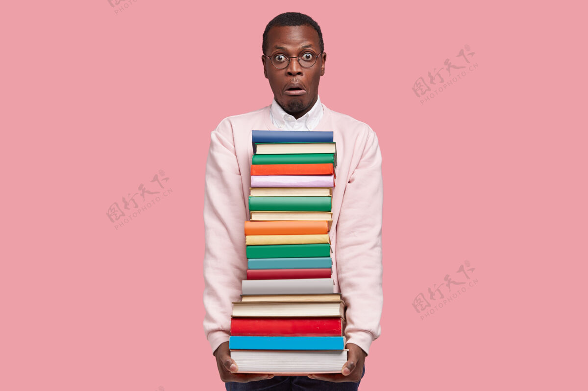 眼镜惊呆了的美国黑人年轻人拿着一大摞书 穿着休闲毛衣 有着惊讶的表情姿势可怕堆