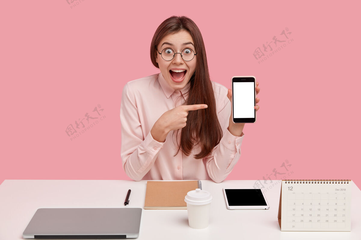设备开心快乐的女人用模拟屏幕指着手机 有着惊艳的表情 完美主义者 力求完美显示在线震惊