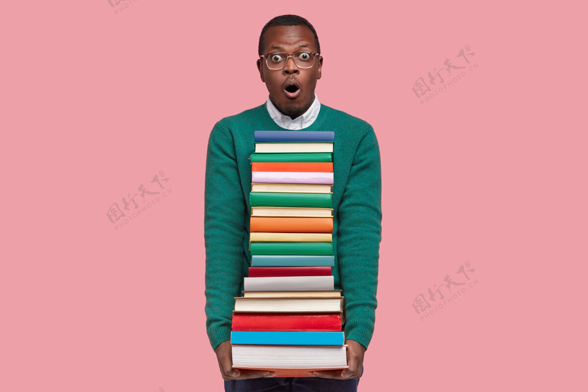 阅读情绪激动的惊讶黑人表情惊恐 拿着一堆教科书 害怕有许多任务要准备 粉红色背景上的模型高中沮丧恐惧