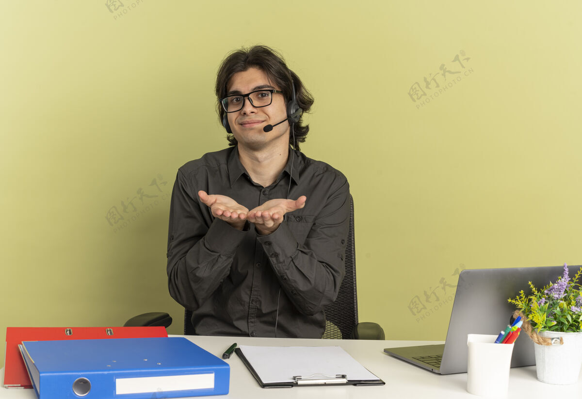 光学戴着耳机 戴着眼镜的年轻上班族坐在办公桌旁 手里拿着笔记本电脑 双手合十复制桌子眼镜