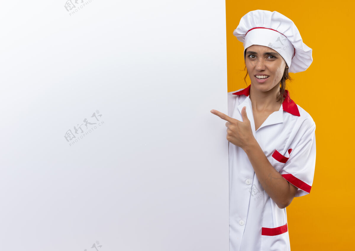 戴着迷茫的年轻女厨师穿着厨师制服 拿着手指头对着白色的墙壁 在隔离的黄色墙壁上留着临摹的空间厨师拿着困惑