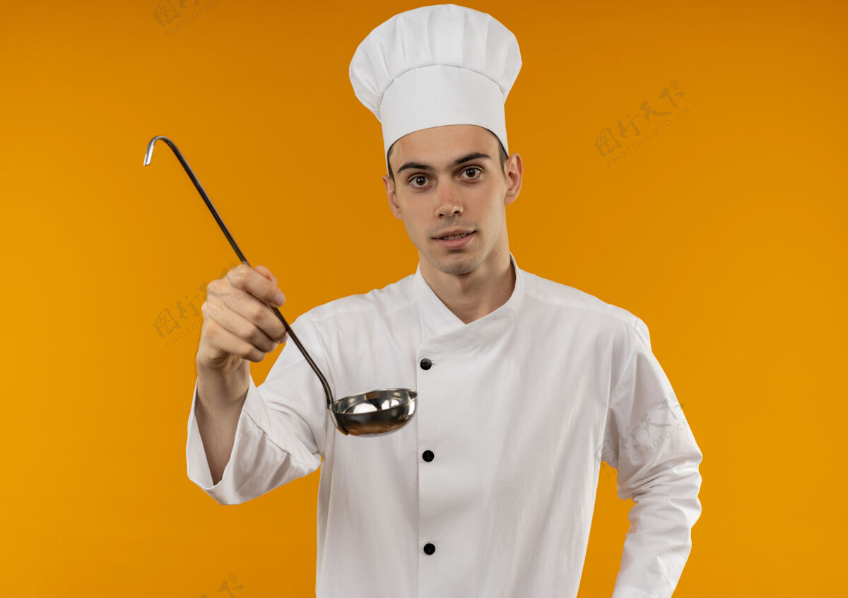 酷年轻的男性穿着厨师制服拿着勺子站在隔离的黄色墙上看抱厨师
