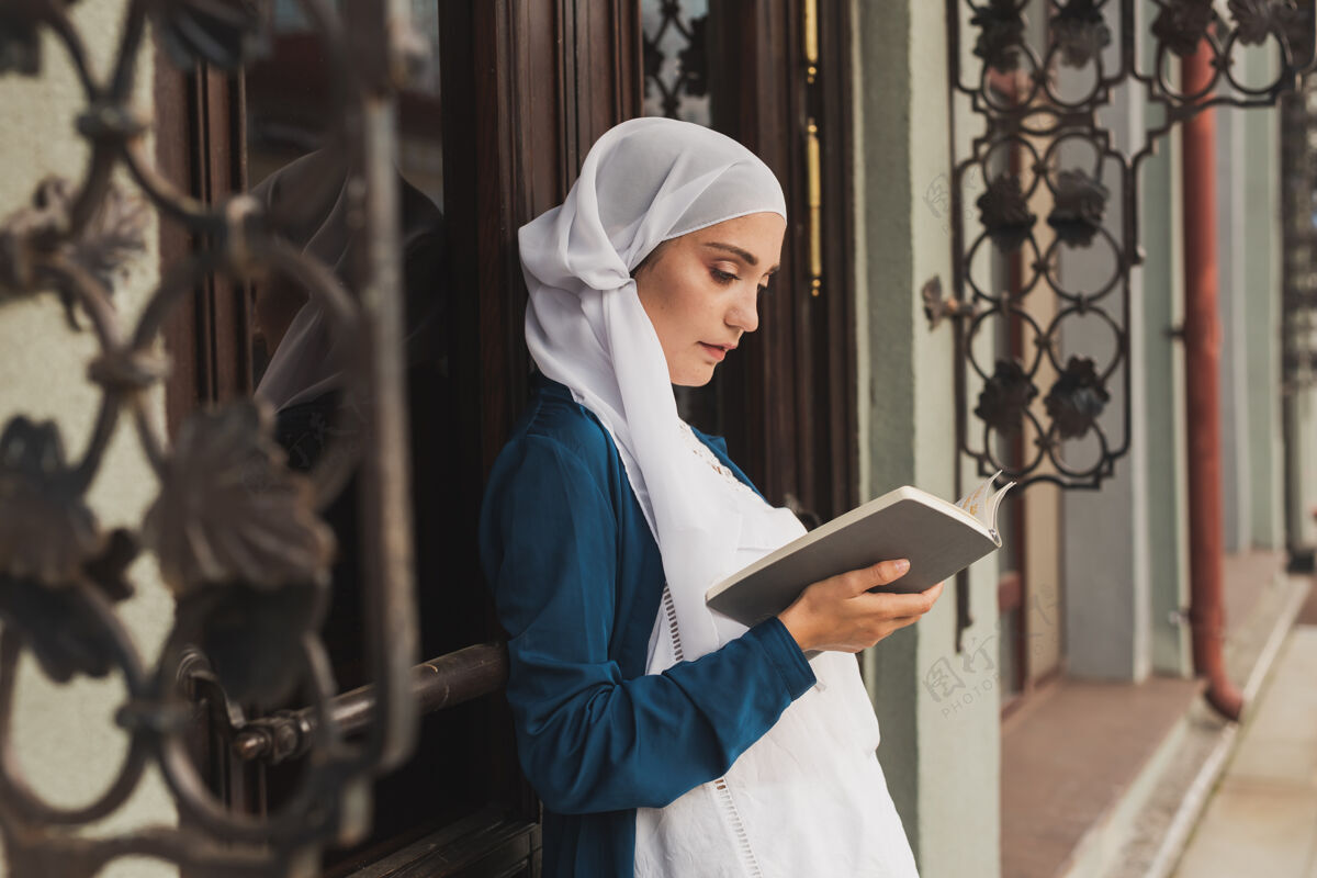 女孩年轻的穆斯林妇女戴着头巾在户外看书的画像教科书家庭作业学者