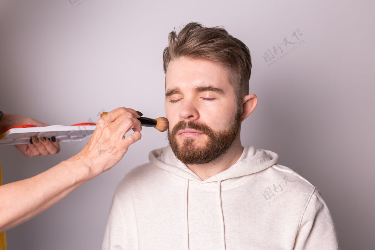 Metrosexual胡子男人正在用刷子给visagist化妆工作工作护理