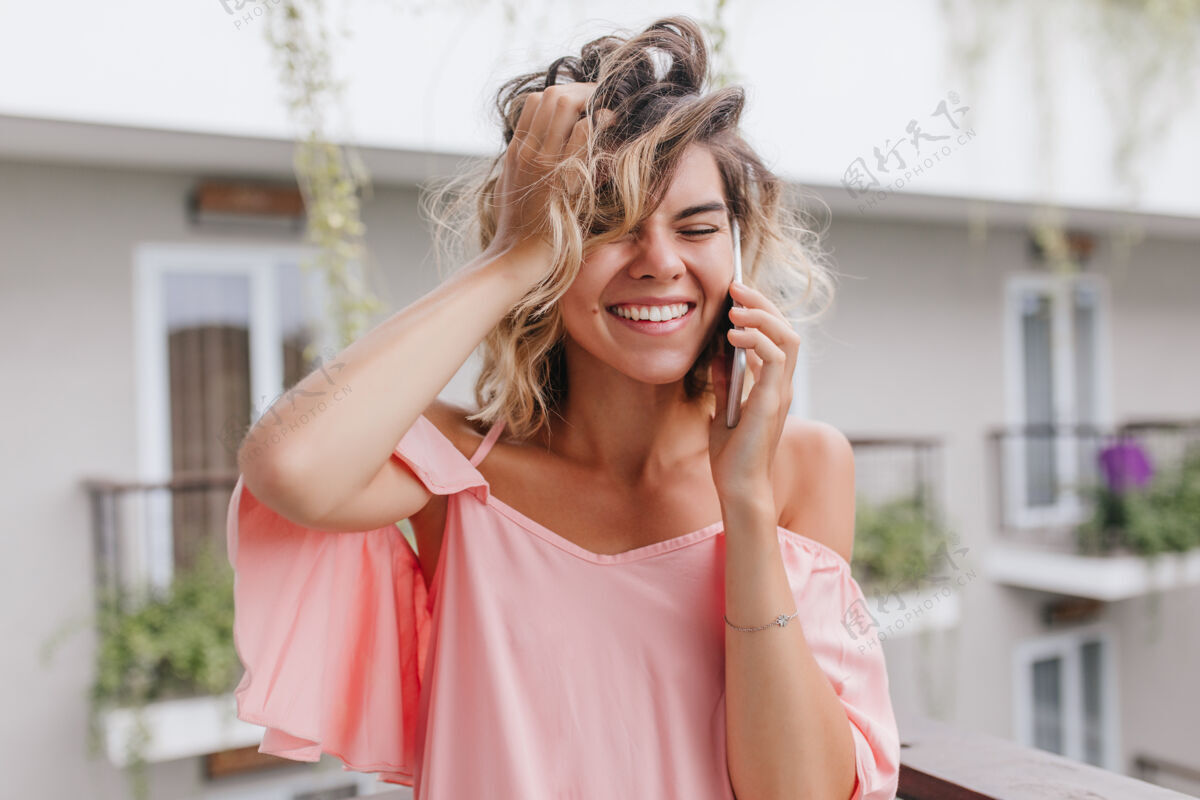 人无忧无虑的金发女郎微笑着讲电话的特写照片迷人的女模特在通话中玩弄头发的户外照片露台情感酒店